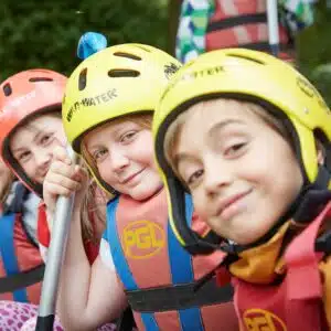 Three children wearing helmets and life jackets, holding a paddle, smiling at the camera with trees in the background.