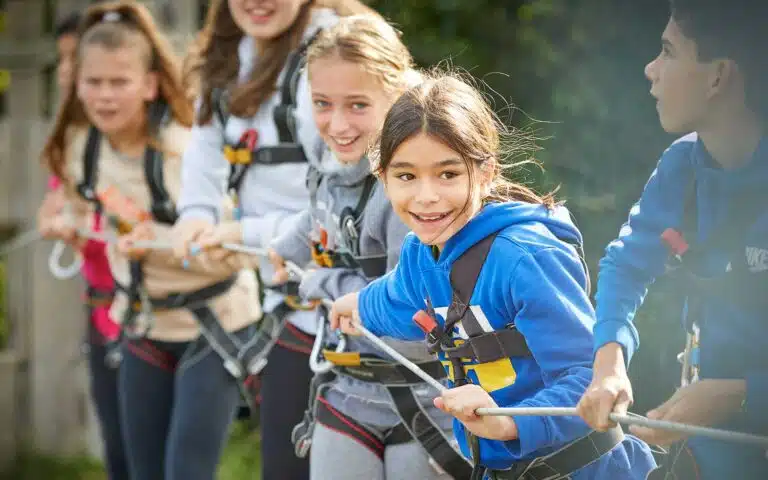 Group of children in harnesses participating in a multi-activity outdoor event, smiling and focused on the tug-of-war game.