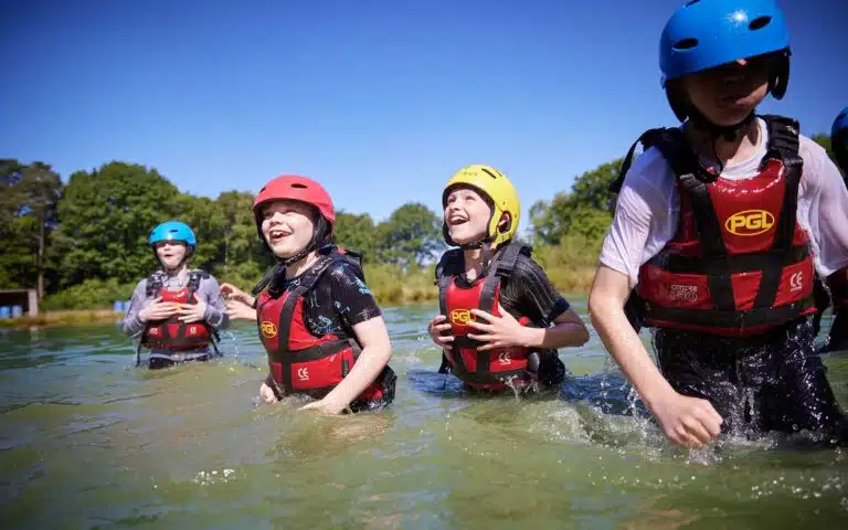 Children with helmets and life jackets laugh and splash in a sunny lake during an outdoor multi-activity water event.