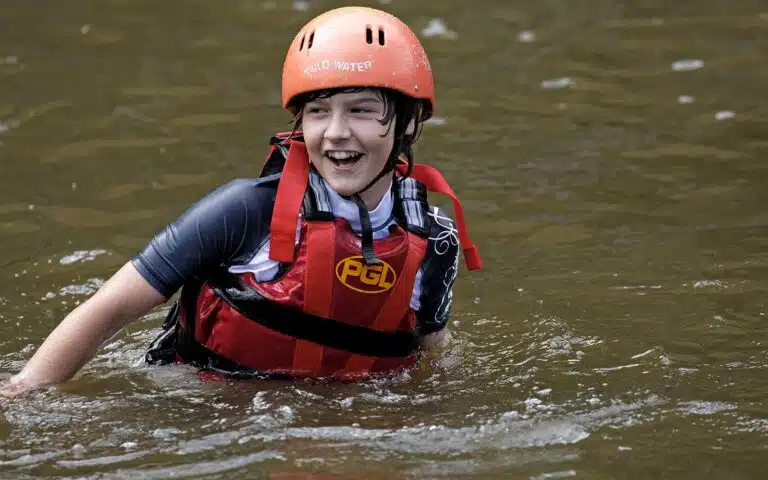 Teenager in a red life vest and orange helmet smiling while participating in a multi-activity adventure, wading through a river.