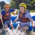 Two people in helmets and life jackets enthusiastically paddling and reacting to splashing water in a raft.