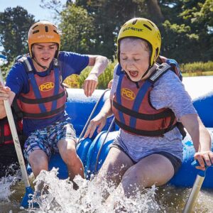 Two people in helmets and life jackets enthusiastically paddling and reacting to splashing water in a raft.