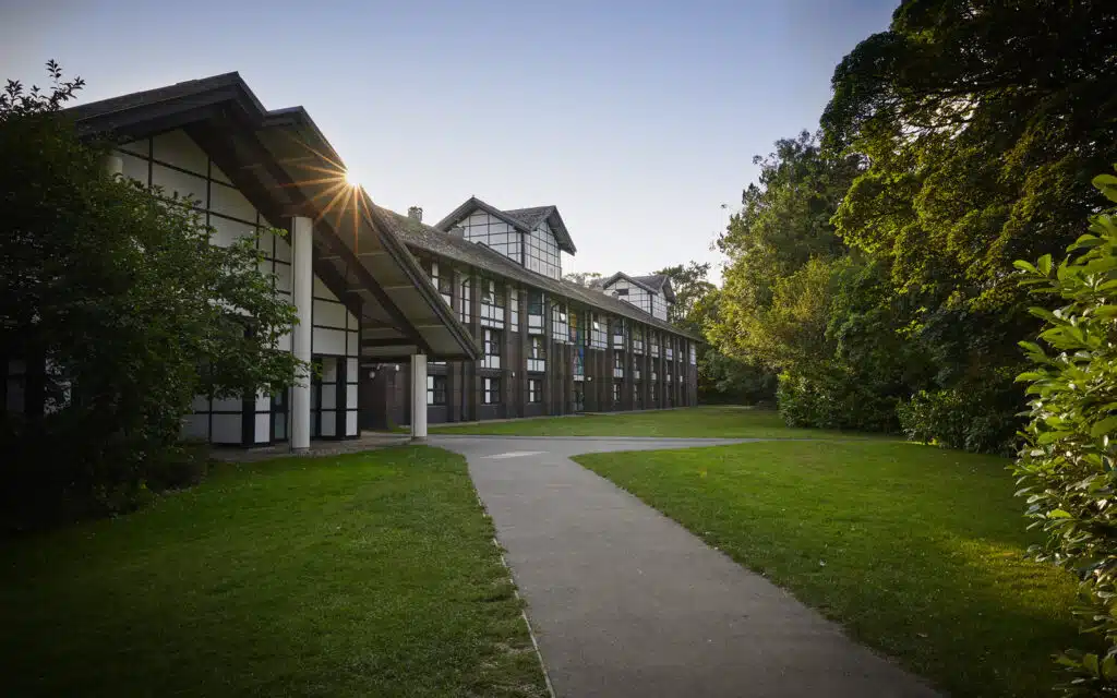 Modern two-story building with large glass windows surrounded by lush greenery, with a pathway leading towards it and the sun setting behind.