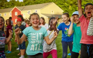 A group of children joyfully running on grass, with a girl in a turquoise shirt leading and laughing, houses in the background.