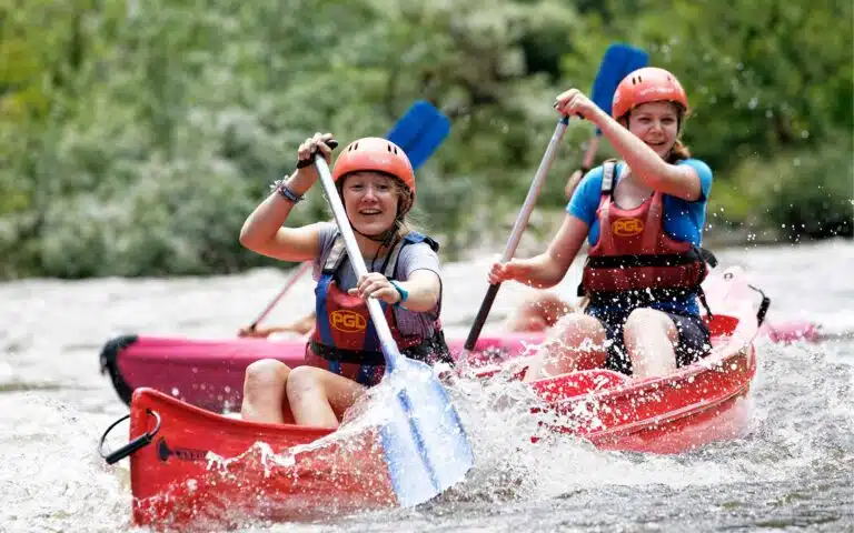 Two people in helmets and life jackets excitedly paddle a red kayak through splashing river waters during a multi-activity adventure.