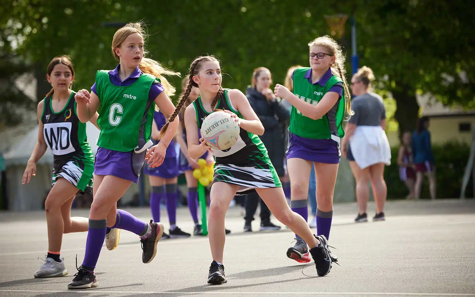 Young girls in green and purple uniforms playing netball on an outdoor court, passing and defending during a daytime game.
