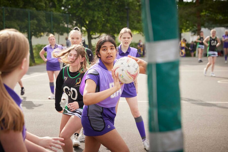 Young female netball players in action on an outdoor court at a PGL Adventure Holidays camp, with one player in purple about to pass the ball.