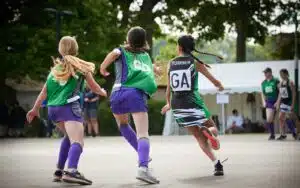 Three female netball players in motion on a court, with one team in green and purple uniforms and the other in green and black.