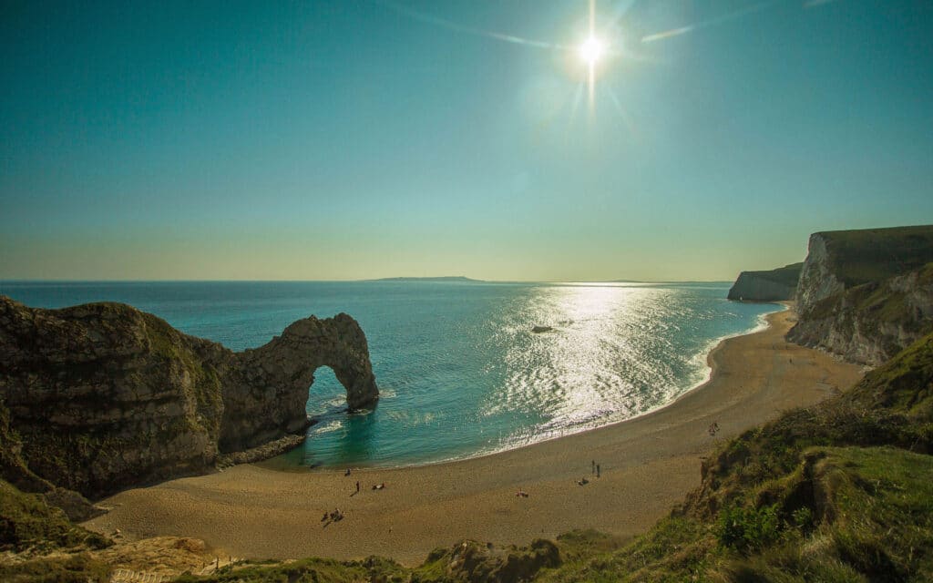 Sun shines above Durdle Door, a natural limestone arch on a sandy beach surrounded by cliffs, with calm sea extending into the horizon.