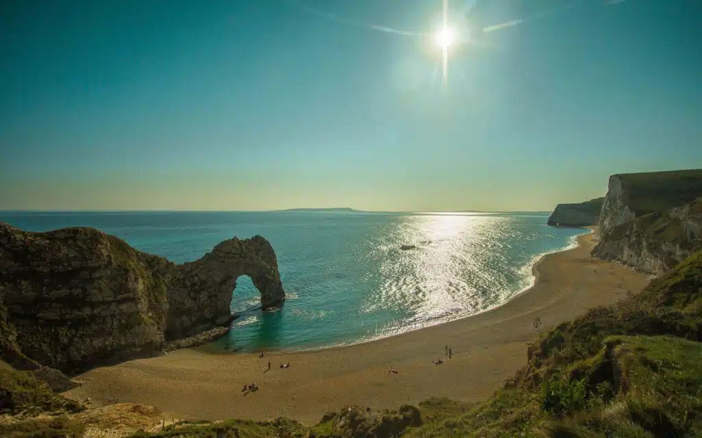 Sun shines above Durdle Door, a natural limestone arch on a sandy beach surrounded by cliffs, with calm sea extending into the horizon.