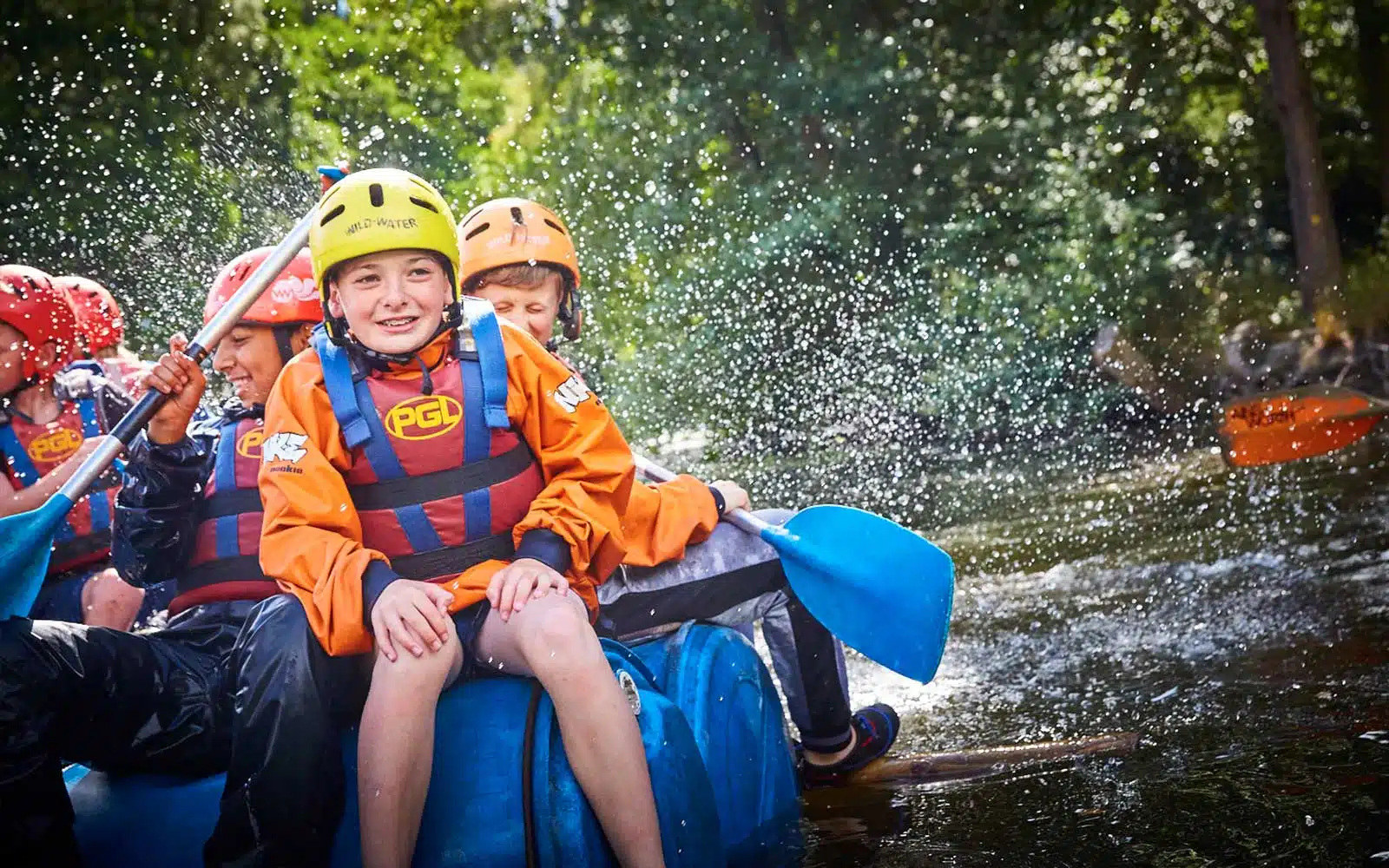 Children in helmets and life jackets laughing while rafting on a river, with water splashing around them.
