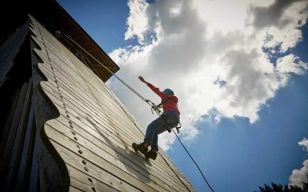 Person climbing a wooden wall with a rope, under a cloudy sky.