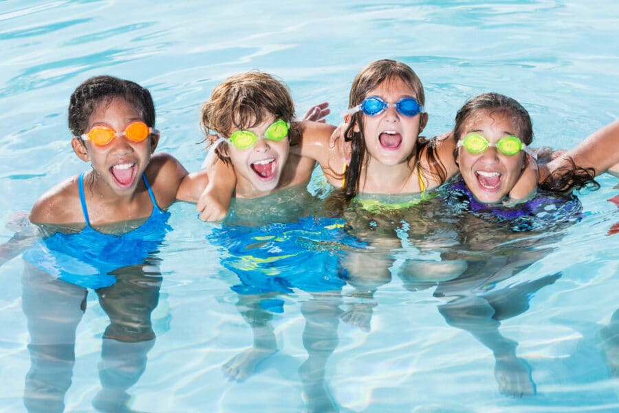 Four children with swim goggles making playful faces in a swimming pool.