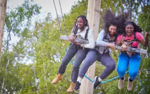 Three joyful women swinging side by side on a high ropes adventure course, secured with safety harnesses.