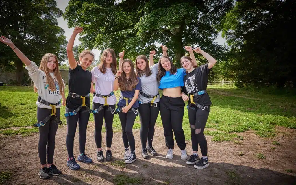 Group of seven young females wearing climbing harnesses, smiling and raising their arms in a sunny park.