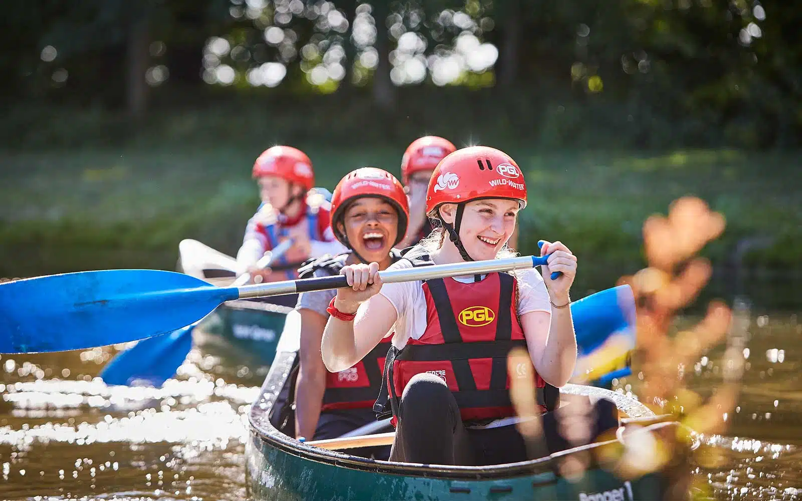 Two people wearing red helmets and life jackets paddling a canoe, with a focus on a smiling woman in the foreground.