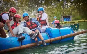 Group of joyful people with helmets and life jackets riding a blue raft on a lake, splashing water around.