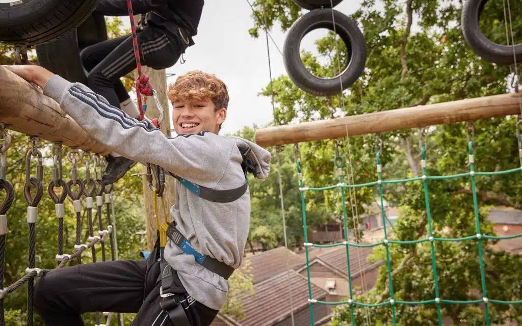Young man in safety harness smiling while navigating a high ropes obstacle course with tire elements and green trees in background.