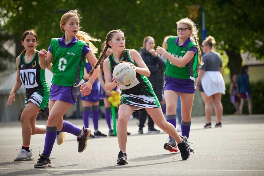 Young girls in green and purple uniforms playing netball on an outdoor court, one in purple dribbling the ball forward.