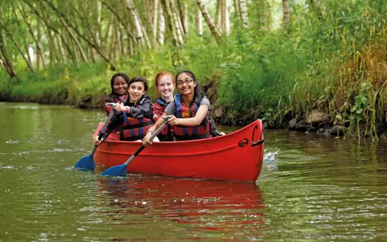 Four children wearing life jackets engage in a multi-activity adventure, paddling a red canoe on a calm, narrow river surrounded by lush greenery.