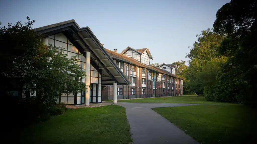 Modern office building with large glass windows and a triangular roof, surrounded by green trees and a path leading up to it.