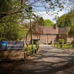 A serene view of Bethany House, a brick building surrounded by lush greenery, with a prominent sign at the driveway entrance.