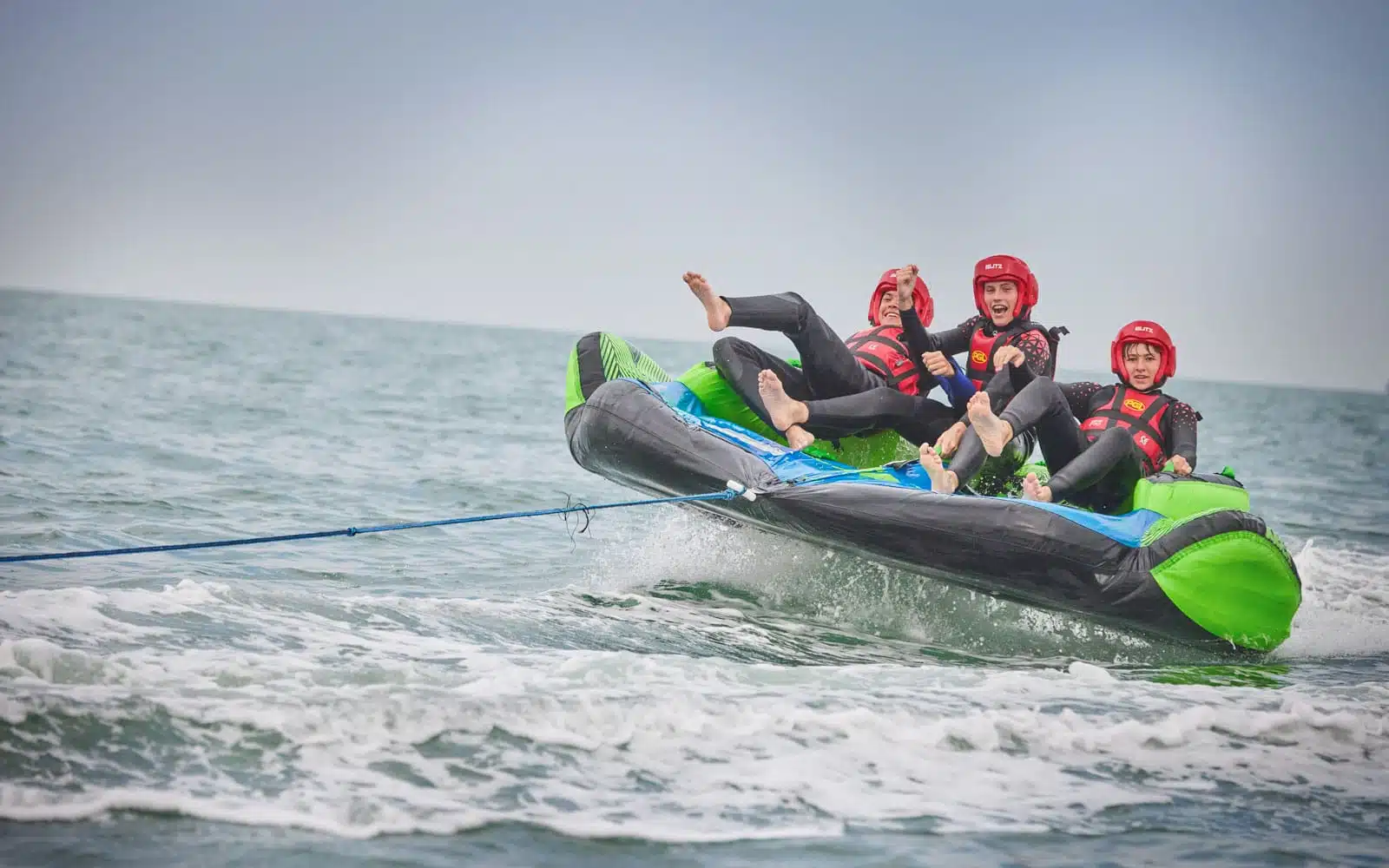 Four people in red helmets and life jackets engage in a multi-activity adventure, riding a green and black inflatable tube being towed by a boat on choppy sea waters.