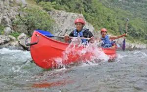 Two people in a red canoe navigate rough river waters, splashing through while paddling energetically during their multi-activity adventure, surrounded by rocky hills.
