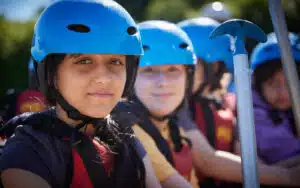 Three young people wearing helmets and life vests, holding a paddle, ready for a water sporting adventure.