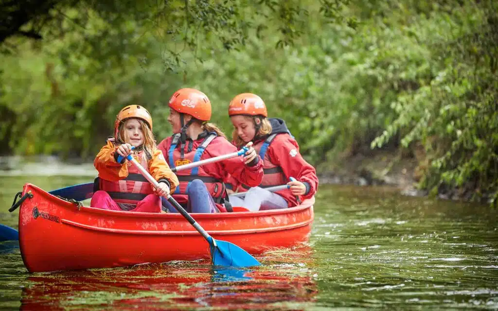 Three individuals in orange helmets and red life vests paddle a red canoe along a river surrounded by greenery, enjoying a multi-activity adventure.