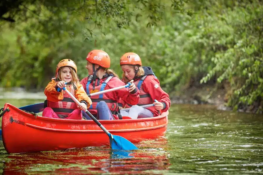 Three individuals in orange helmets and red life vests paddle a red canoe along a river surrounded by greenery, enjoying a multi-activity adventure.