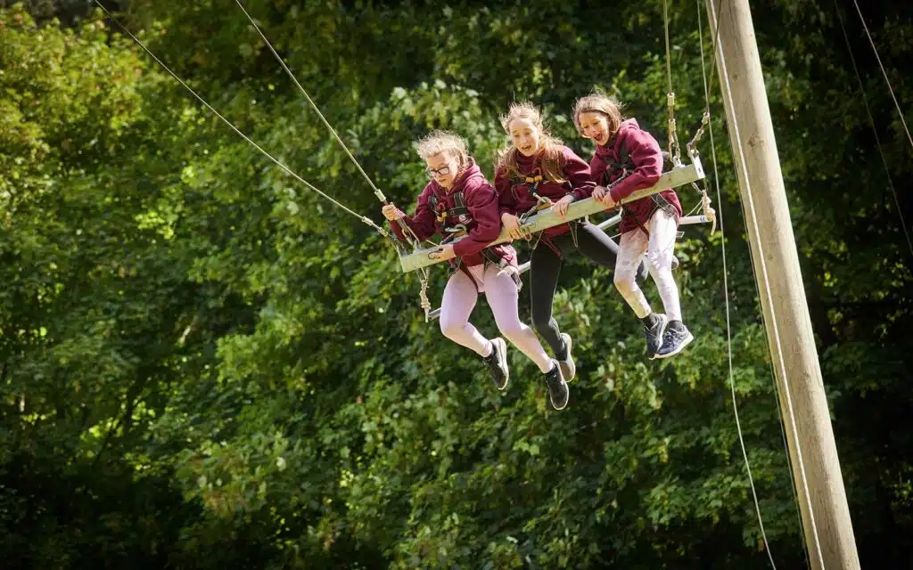 Three people having fun on a multi-activity swing ride at an outdoor adventure park, surrounded by trees.