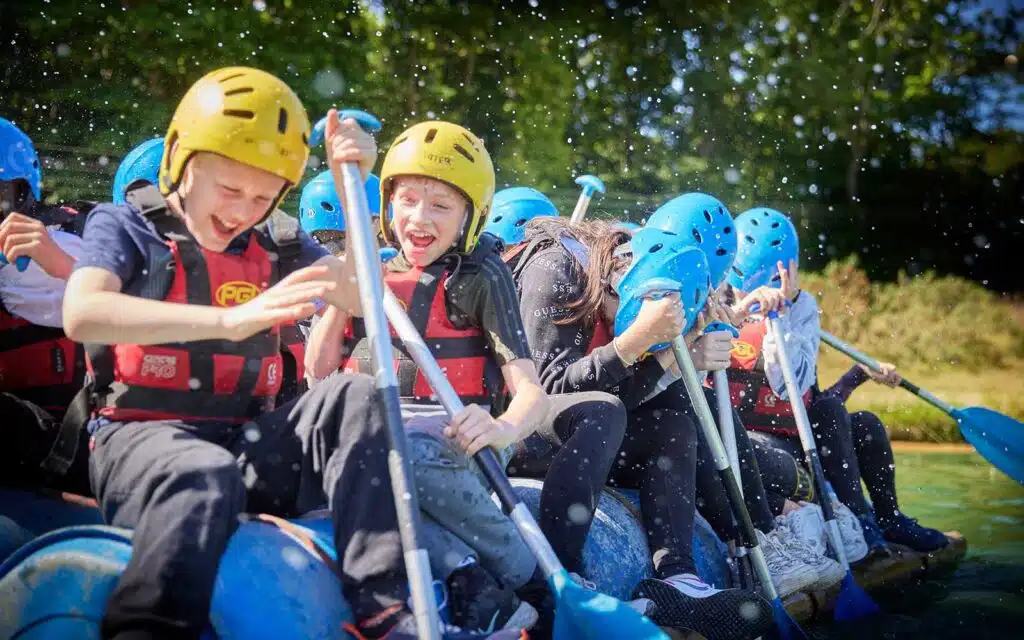 A group of children wearing helmets and life jackets enthusiastically paddling in a multi-activity raft on a sunny day.