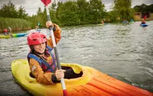 A young child wearing a red helmet and life vest smiles while paddling an orange kayak on a tranquil lake during a multi-activity day.