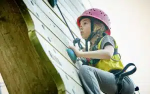 A young girl wearing a helmet and harness climbs an outdoor rock wall, looking focused and determined.