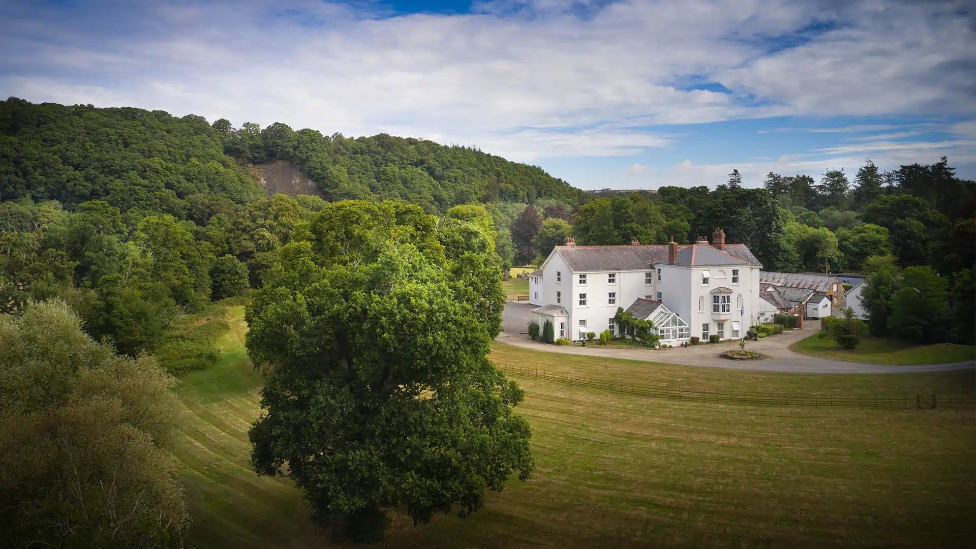 Aerial view of a large white house with multiple annexes, surrounded by green lawns and dense trees under a partly cloudy sky.