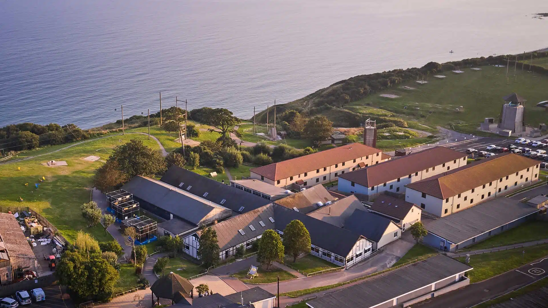 Aerial view of a coastal area with buildings, a lighthouse, and a green park at sunset.