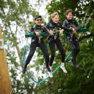 Three boys wearing harnesses joyfully swing from a zip line amidst trees, showing excitement and enjoyment.
