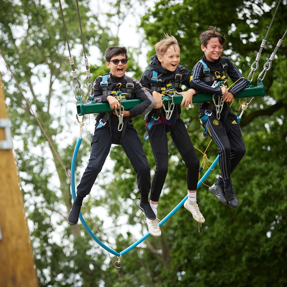 Three boys wearing harnesses joyfully swing from a zip line amidst trees, showing excitement and enjoyment.
