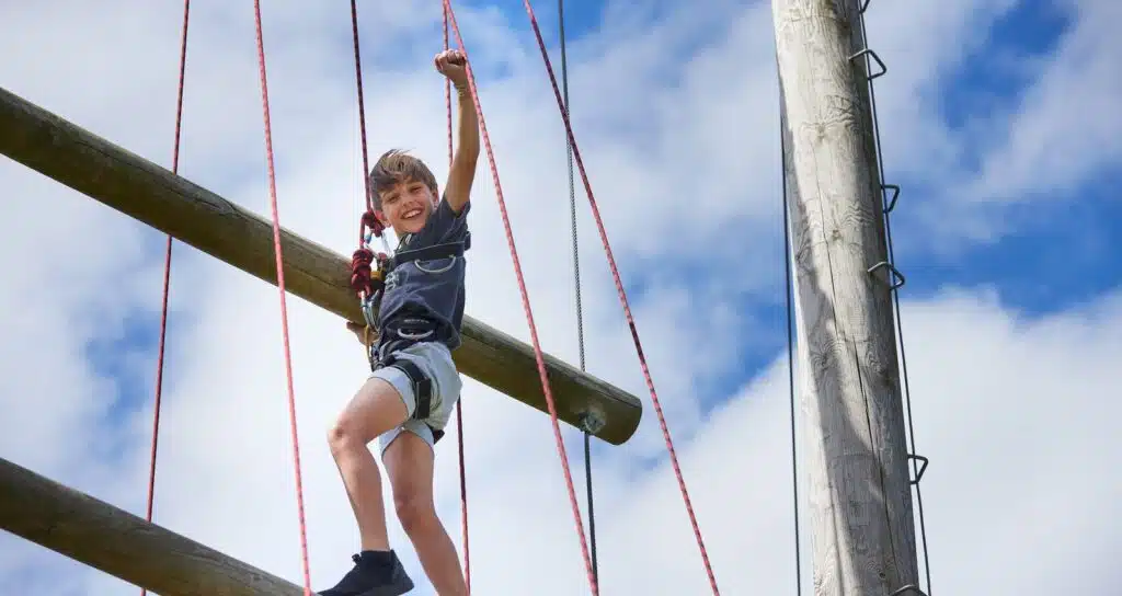 A young boy with a joyful expression climbs on a high rope course against a clear blue sky.