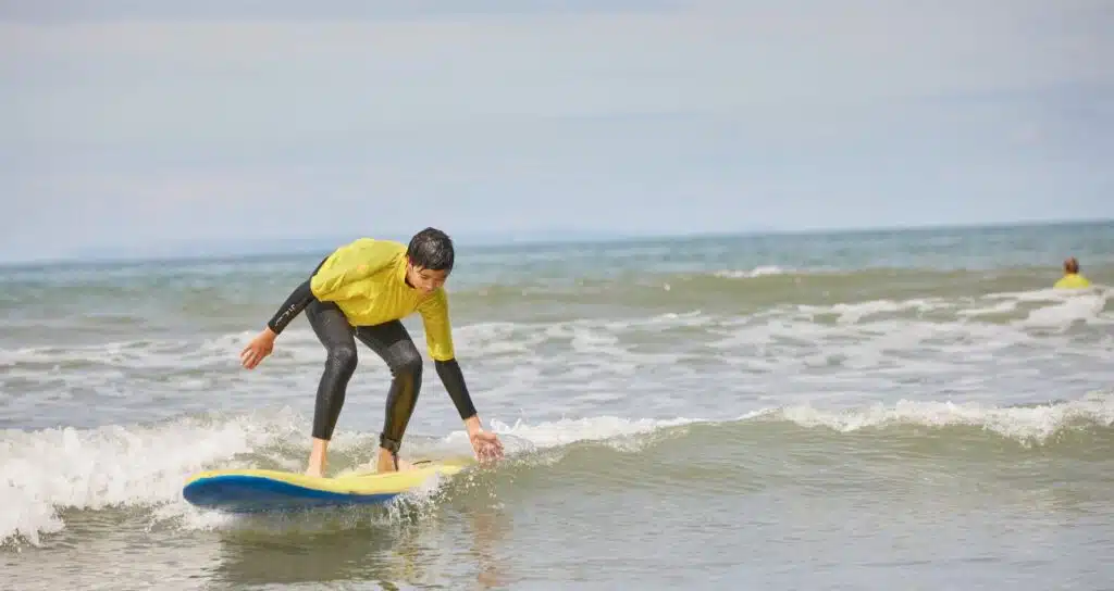 Young person engaged in physical activity benefits for kids, wearing a yellow rash guard and black wetsuit, surfing a small wave, with clear sky and distant swimmer in the background.