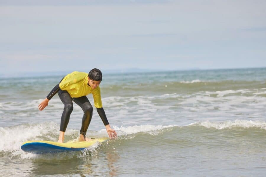Young person engaged in physical activity benefits for kids, wearing a yellow rash guard and black wetsuit, surfing a small wave, with clear sky and distant swimmer in the background.