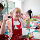 Young girl in a cooking class, smiling with flour on her hands, wearing a red apron and a chef's hat, standing in a kitchen at Pony Camp Trekker - Tregoyd House on Jul 27, 2024.
