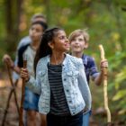 A young girl leading a group of children hiking in a forest, smiling and looking to the side, holding a Bushcraft walking stick.