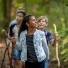 A young girl leading a group of children hiking in a forest, smiling and looking to the side, holding a Bushcraft walking stick.