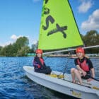 Two people wearing life jackets and helmets sailing a Pony Camp Trekker boat with a green sail on a lake at Tregoyd House - Jul 27, 2024.