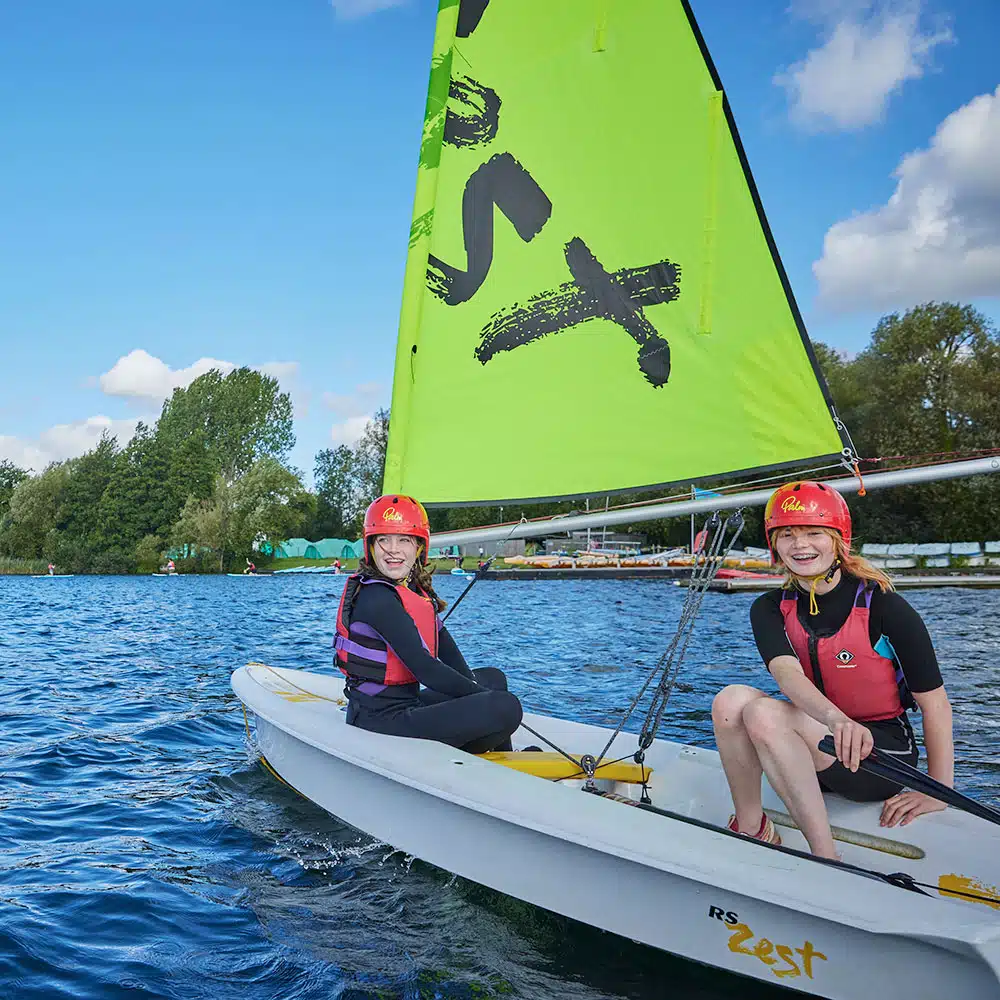 Two people wearing life jackets and helmets sailing a Pony Camp Trekker boat with a green sail on a lake at Tregoyd House - Jul 27, 2024.