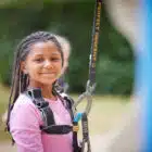 A young girl with braided hair smiles, wearing a Multi-Activity, Marchants Hill - May 28, 2024 harness and holding a climbing rope, ready for an adventure.
