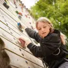 A young girl wearing a Pony Camp Trekker climbs an outdoor climbing wall, reaching for a handhold, with trees in the background at Tregoyd House on Jul 27, 2024.