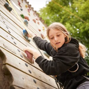 A young girl wearing a Pony Camp Trekker climbs an outdoor climbing wall, reaching for a handhold, with trees in the background at Tregoyd House on Jul 27, 2024.
